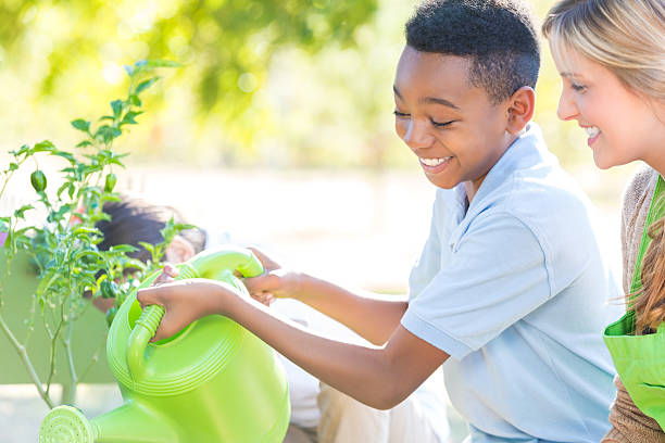 African American elementary age little boy is smiling while using a watering can to water vegetable plants in school garden. Mid adult Caucasian woman is teaching class to diverse private school students, outdoors in school garden. Students are learning science with hands on experience. They are wearing uniforms.